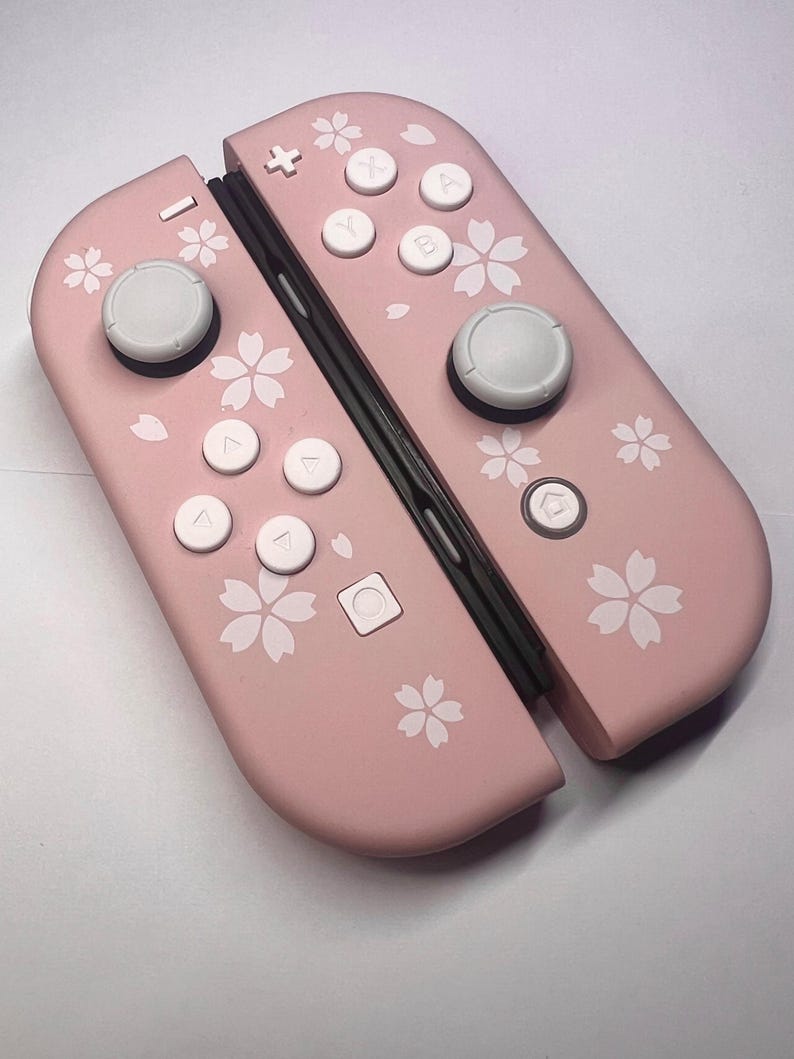 以下が含まれることがあります： ピンク色のNintendo Switch Joy-Conコントローラー。白いボタンと花の模様が特徴です。 コントローラーは明るいピンク色で、白い桜の花のパターンが施されています。 ボタンは白で、ジョイスティックはグレーです。