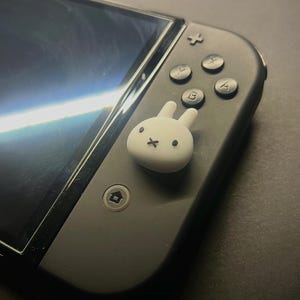 May include: A black Nintendo Switch controller with a white bunny-shaped thumb grip. The controller has black buttons with white lettering and a black screen. The bunny grip has a simple face design.