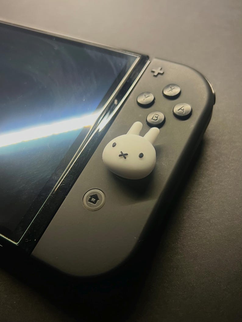 May include: A black Nintendo Switch controller with a white bunny-shaped thumb grip. The controller has black buttons with white lettering and a black screen. The bunny grip has a simple face design.