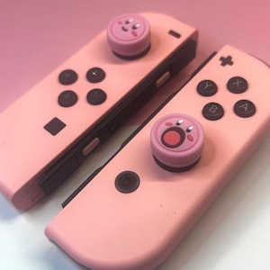 May include: Two pink Nintendo Switch Joy-Cons with black buttons and a pink character-themed thumbstick cover. The Joy-Cons are set against a pink background.
