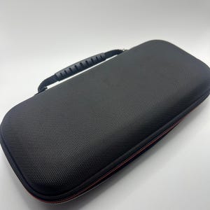 May include: A rectangular black carrying case with a textured, hardshell exterior and a black plastic handle attached to the top. The case features a zipper closure with a thin red accent line running along the perimeter of the storage unit.