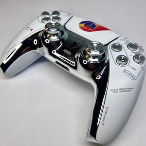May include: White and chrome video game controller with a colorful design on the center. The controller has multiple buttons and joysticks, with text that reads "NOVACS REMOTE MANUAL PILOT CONTROLLER" and "GRIP HERE".