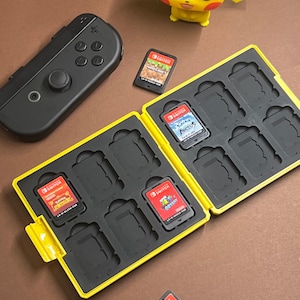 Könnte beinhalten: Eine gelbe Switch-Spielkassettenhülle mit schwarzem Innenraum, die mehrere Spielkassetten enthält. Ein schwarzer Switch-Controller und eine gelbe Pikachu-Figur sind ebenfalls sichtbar. Die Kassetten sind mit Spieltiteln beschriftet.