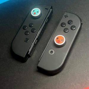 May include: Two black Nintendo Switch Joy-Con controllers. Each controller has a circular button with a cartoon face. One button is light blue with a green face, and the other is orange with a red face. The controllers are on a black surface.