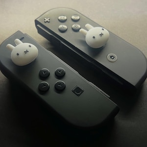 May include: Two black game controllers with white bunny-shaped thumb grips. The controllers have black buttons and directional pads. The bunny grips add a cute, playful touch to the gaming accessories.