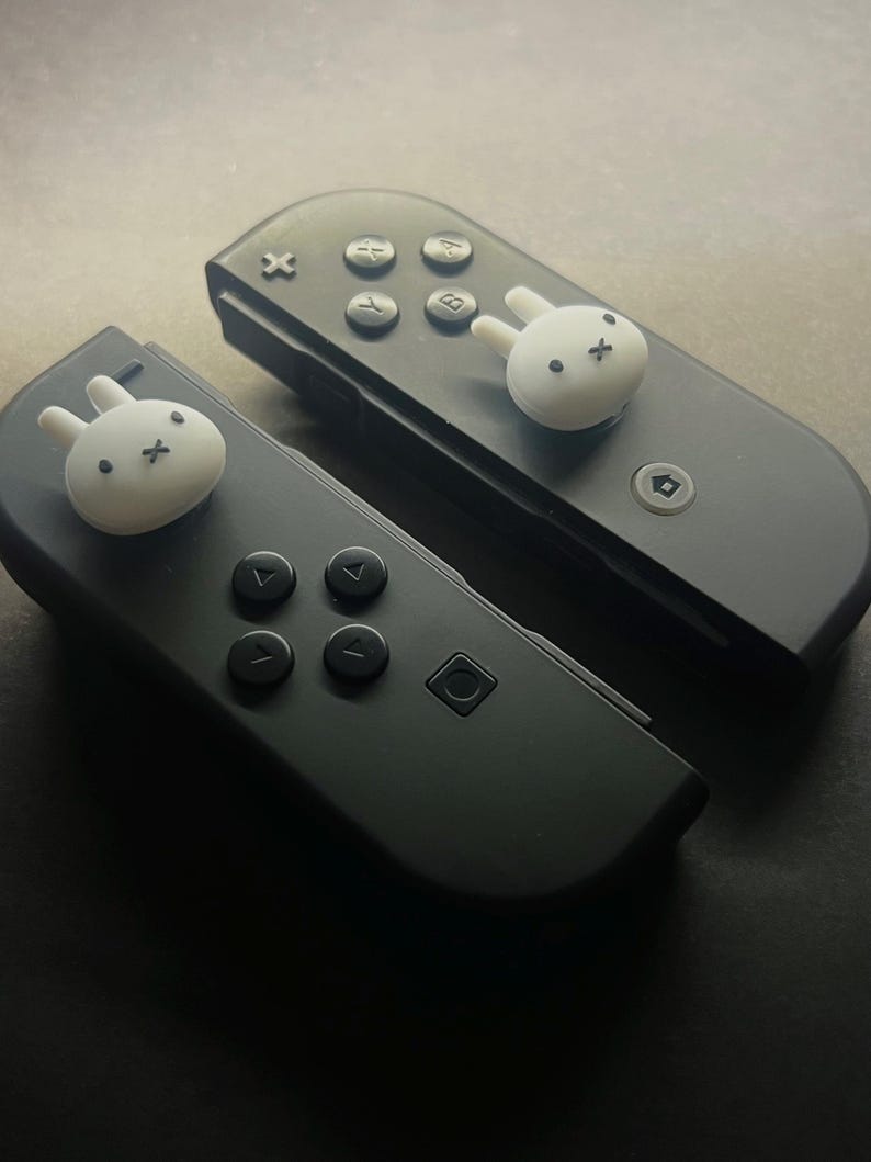May include: Two black game controllers with white bunny-shaped thumb grips. The controllers have black buttons and directional pads. The bunny grips add a cute, playful touch to the gaming accessories.