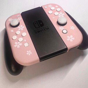 以下が含まれることがあります： ピンク色のNintendo Switch Joy-Conコントローラー。白いボタンと黒い中央部があります。ピンクの部分には白い桜の花模様が施されています。白いジョイスティックと黒いグリップが付いています。