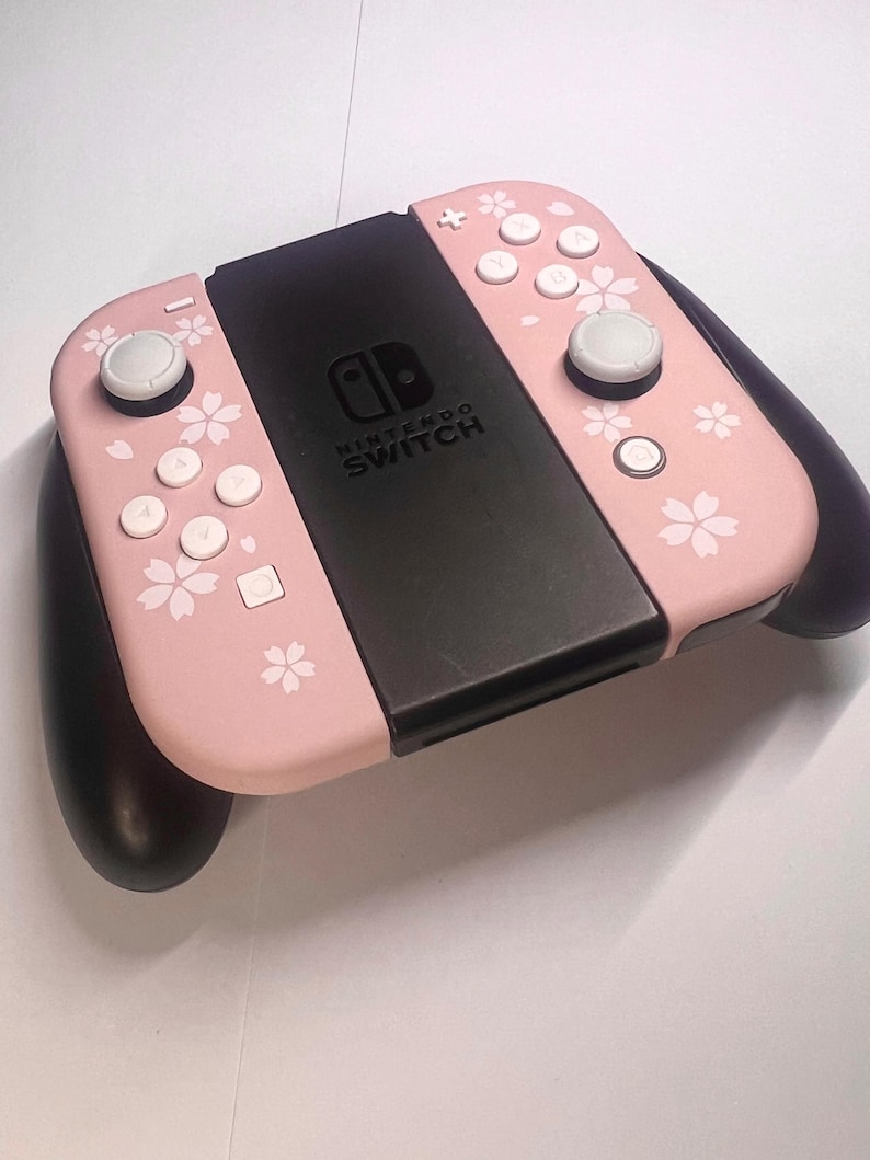 以下が含まれることがあります： ピンク色のNintendo Switch Joy-Conコントローラー。白いボタンと黒い中央部があります。ピンクの部分には白い桜の花模様が施されています。白いジョイスティックと黒いグリップが付いています。