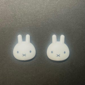 May include: Two white bunny-shaped decorative objects with black eyes and a black 'X' for a mouth. The objects have two upright ears and are set against a dark grey background.