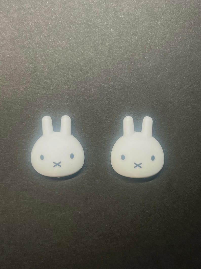 May include: Two white bunny-shaped decorative objects with black eyes and a black 'X' for a mouth. The objects have two upright ears and are set against a dark grey background.