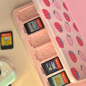 May include: A pink Nintendo Switch game cartridge case decorated with strawberries. The case is open, displaying several game cartridges, including "Pokémon Arceus", "Zelda", and "Animal Crossing". A single game cartridge is visible to the left.