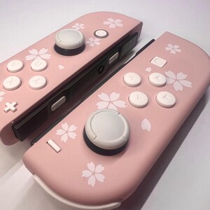 以下が含まれることがあります： 白ボタンとジョイスティックが付いたピンク色のゲームコントローラー2つ。 コントローラーは白い花の模様で装飾されています。 コントローラーはゲーム機用に設計されています。