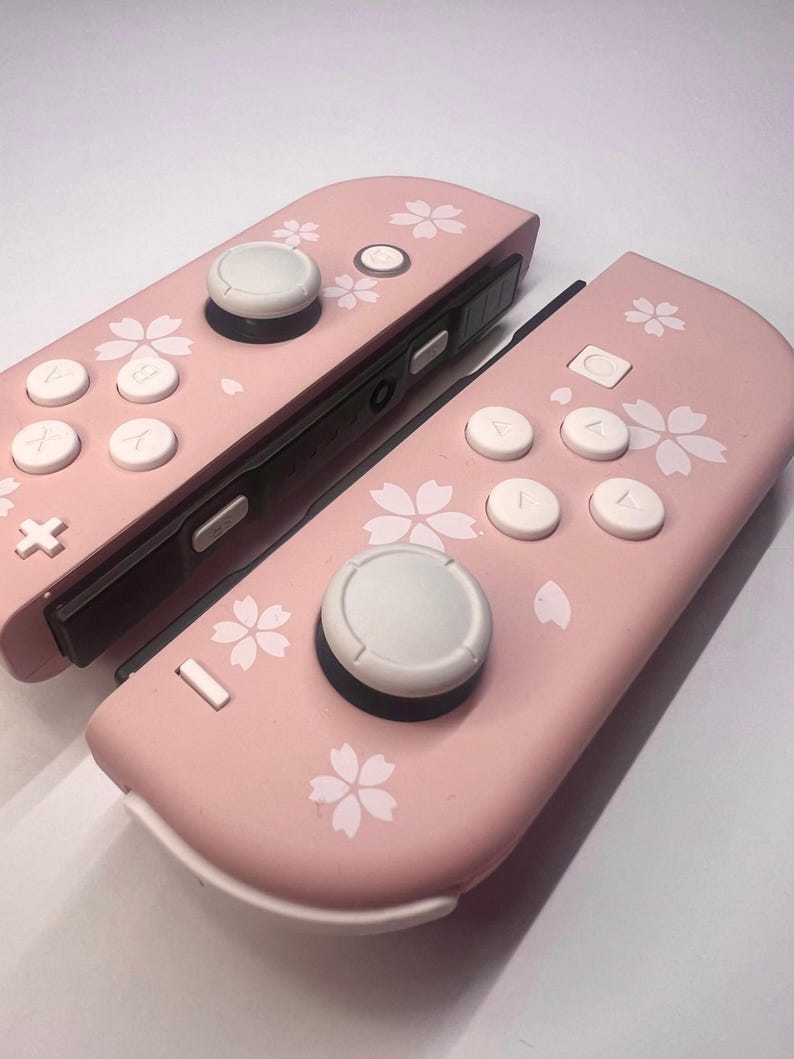 以下が含まれることがあります： 白ボタンとジョイスティックが付いたピンク色のゲームコントローラー2つ。 コントローラーは白い花の模様で装飾されています。 コントローラーはゲーム機用に設計されています。