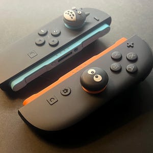 以下が含まれることがあります： 2つの黒いゲームコントローラー。親指グリップは漫画のキャラクターです。1つのコントローラーは水色、もう1つはオレンジ色です。コントローラーには黒いボタンと方向パッドがあります。