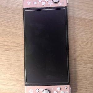 以下が含まれることがあります： 黒い画面のピンクと白のNintendo Switchコンソール。コントローラーはピンク色で、白い花の模様とボタンが付いています。コンソールは木製の表面にあります。