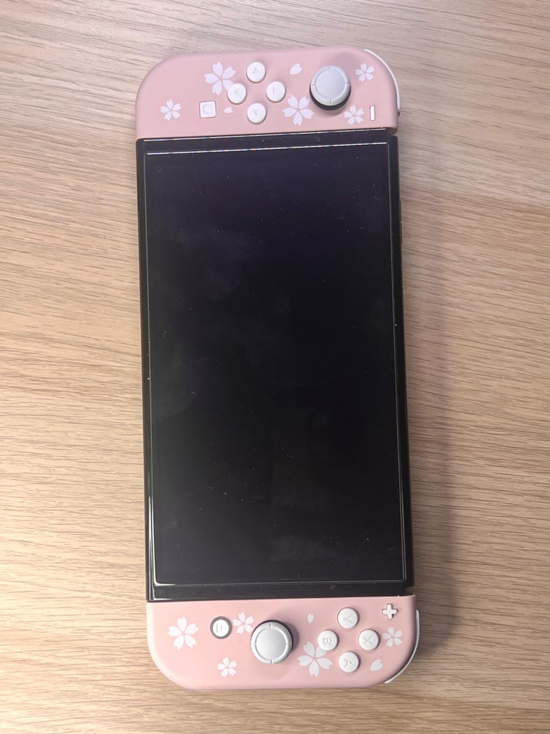 以下が含まれることがあります： 黒い画面のピンクと白のNintendo Switchコンソール。コントローラーはピンク色で、白い花の模様とボタンが付いています。コンソールは木製の表面にあります。