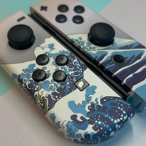 May include: A pair of white and gray game controllers with a blue wave pattern. The controllers have black buttons and joysticks. The wave design is a prominent feature, adding a decorative element to the gaming accessories.