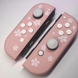 以下が含まれることがあります： 白ボタンと花の模様が付いた、2つのピンク色のゲームコントローラー。 コントローラーは、白い花の模様が入った明るいピンク色のベースカラーです。 ボタンは白で、ジョイスティックはグレーです。