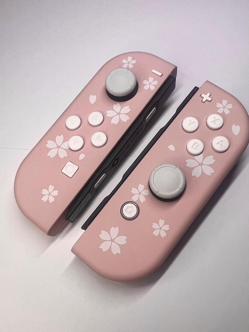 以下が含まれることがあります： 白ボタンと花の模様が付いた、2つのピンク色のゲームコントローラー。 コントローラーは、白い花の模様が入った明るいピンク色のベースカラーです。 ボタンは白で、ジョイスティックはグレーです。