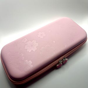 Könnte beinhalten: Ein rechteckiges, hellrosa Hartschalenetui mit einem geprägten Kirschblütenmuster auf der Oberseite. Das Etui wird mit einem passenden rosa Reißverschluss verschlossen, der zwei kleine, blütenförmige Zipper für einfaches Öffnen besitzt.