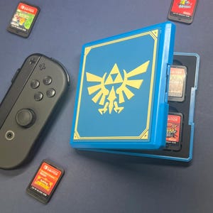 Może przedstawiać: Niebieskie etui na kartridże do gier ze złotym emblematem i czarnym kontrolerem. Widocznych jest kilka kartridży do gier, wraz z etui. Etui jest otwarte, ukazując kartridże do gier w środku. Etui ma złotą obwódkę.