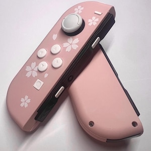 以下が含まれることがあります： ピンク色のゲームコントローラー2つ。白い花の模様があり、白いボタンと灰色のジョイスティックが付いています。ゲーム機用に設計されています。
