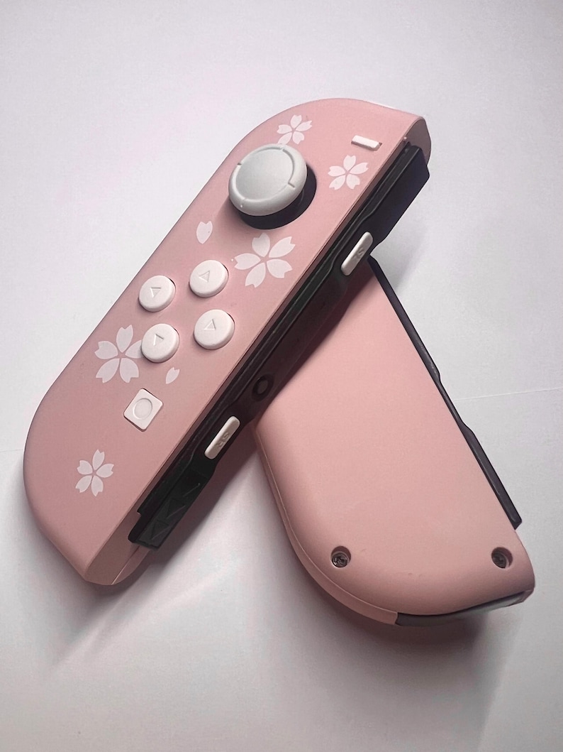 以下が含まれることがあります： ピンク色のゲームコントローラー2つ。白い花の模様があり、白いボタンと灰色のジョイスティックが付いています。ゲーム機用に設計されています。