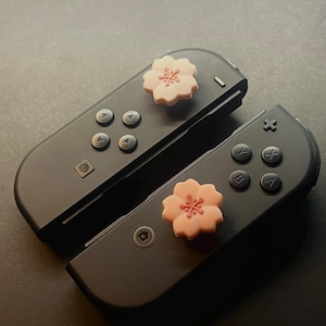 Può includere: Due controller di gioco neri con impugnature per pollice a forma di fiore rosa. I controller sono angolati l'uno verso l'altro, con le impugnature a fiore sulla parte superiore di ogni controller. Lo sfondo è una superficie grigio scuro.