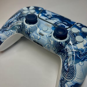 May include: A wireless video game controller featuring a glossy white finish adorned with a detailed blue and white dragon and wave pattern. The device includes two dark blue textured analogue sticks, light blue directional buttons, and a central touchpad.