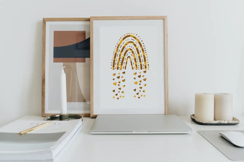 May include: A framed art print featuring a gold and brown rainbow design with heart-shaped accents. The artwork is displayed on a white desk alongside a laptop, candles, and other decorative items. The print is in a wooden frame.