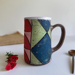 Pode incluir: Uma caneca de cerâmica artesanal com um padrão geométrico em vermelho, rosa, verde e azul. A caneca tem uma alça marrom e uma borda branca. Uma colher de madeira e flores vermelhas estão ao lado da caneca.