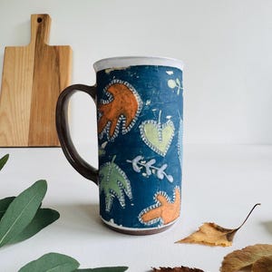 Könnte beinhalten: Eine hohe Keramiktasse mit dunkelblauem Hintergrund und braunem Henkel. Die Tasse ist mit orangefarbenen, grünen und gelben Blattmotiven verziert. Der Rand der Tasse ist weiß. Ein Holzschneidebrett befindet sich im Hintergrund.