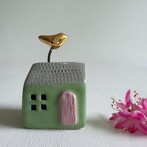 May include: A ceramic house figurine with a green body, gray roof, and pink door. A gold-colored bird sits atop the roof. The house has four black square windows. A pink flower is in the background.
