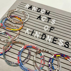 May include: A collection of colorful beaded bracelets stacked on a gray wooden surface with the text "RANDOM ACTS OF KINDNESS" displayed on the background.