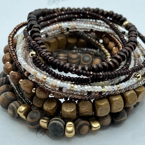 May include: A stack of wooden and beaded bracelets in brown, gold, and white. The bracelets are layered on top of each other, creating a unique and stylish look.