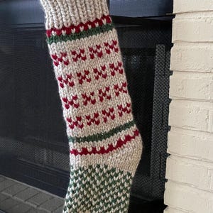 May include: A knitted Christmas stocking with a white background and red and green geometric patterns. The stocking is hanging on a black metal fireplace screen.