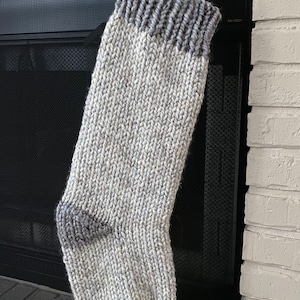 May include: A white and gray knitted Christmas stocking with a gray cuff hanging on a black metal fireplace screen.