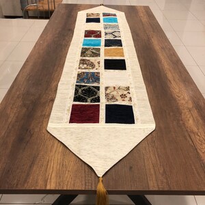 Puede incluir: Un camino de mesa blanco con un patrón geométrico de cuadrados de tela de colores. El camino de mesa tiene un borla en el extremo y es perfecto para agregar un toque de color y estilo a cualquier mesa.