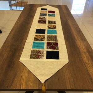 Könnte beinhalten: Ein weißer Tischläufer mit einem Patchwork-Muster aus bunten Quadraten. Die Quadrate zeigen verschiedene Muster und Farben, darunter Blau, Grün, Rot und Gold. Der Läufer hat einen Fransenrand und Quasten an den Enden.
