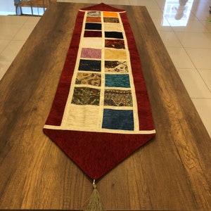 Puede incluir: Un camino de mesa rojo con un diseño de patchwork decorativo que presenta varios colores y patrones. El camino de mesa tiene un borla en el extremo.