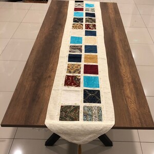 Puede incluir: Un camino de mesa blanco con un diseño de patchwork de cuadrados coloridos. Los cuadrados presentan varios patrones y texturas, incluyendo diseños florales, geométricos y abstractos. El camino de mesa tiene un borde con flecos y una borla en el extremo.