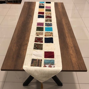 Puede incluir: Un camino de mesa blanco con un diseño de patchwork de cuadrados coloridos. Los cuadrados presentan varios patrones y colores, incluyendo rojo, azul, verde y dorado. El camino de mesa está sobre una mesa de madera.