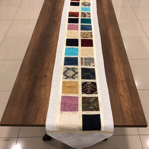 Puede incluir: Un camino de mesa blanco con un patrón decorativo de cuadrados de tela de colores. El camino de mesa tiene un borde de flecos y una borla en el extremo.