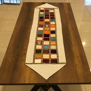 May include: A white table runner with a colorful patchwork design. The runner features a variety of colors, including red, blue, green, yellow, and orange. The runner is trimmed with a gold braid and has a tassel at the end.