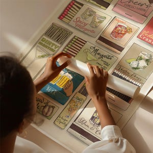 May include: A person rolling out a large poster featuring illustrations of various cocktails. The poster includes images of a Dirty Martini, Margarita, Espresso Martini, and other drinks. The poster has a light background and colorful illustrations.