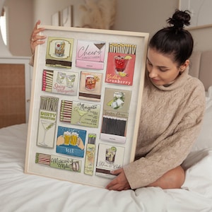 May include: A framed print featuring illustrations of various cocktail matchboxes. The artwork includes images of drinks like an Old Fashioned, Espresso Martini, Vodka Cranberry, and Margarita. The print is held by a person.