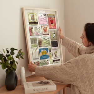 May include: A framed print featuring vintage-style matchbox art. The artwork showcases various cocktail illustrations, including a margarita, vodka cranberry, and beer. The frame is a light color, and the overall theme is bar decor.