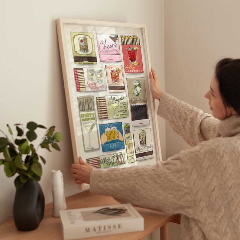 May include: A framed print featuring vintage-style matchbox art. The artwork showcases various cocktail illustrations, including a margarita, vodka cranberry, and beer. The frame is a light color, and the overall theme is bar decor.
