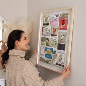 May include: A framed art print featuring colorful illustrations of cocktail matchbooks. The artwork includes images of various drinks like vodka cranberry, margarita, and Moscow mule. The frame is a light wood color, and the print is displayed on a neutral wall.