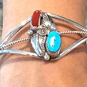 May include: Silver cuff bracelet with a central design featuring a red stone, a turquoise stone, and silver feather accents. The bracelet has a split band design and is worn on a wrist.